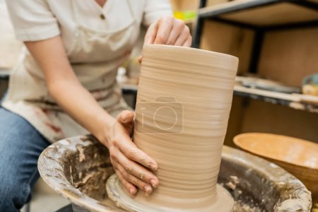 Vista recortada de la artesana borrosa en delantal creando jarrón de arcilla en la rueda de cerámica en el taller