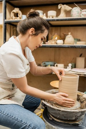 Foto de Vista lateral de la artesana en delantal trabajando con jarrón de arcilla sobre rueda de cerámica en estudio en taller - Imagen libre de derechos