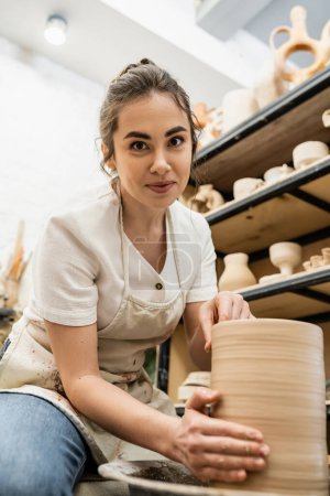 Morena artesana en delantal mirando a la cámara y haciendo jarrón de arcilla en la rueda de cerámica en el taller