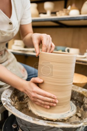 Cropped view of craftswoman in blurred apron making clay vase on pottery wheel in ceramic studio