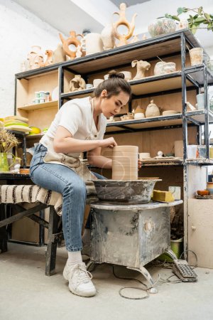 Téléchargez les photos : Brune femme potier en tablier et vêtements de travail faisant vase d'argile sur roue de poterie en atelier de céramique - en image libre de droit