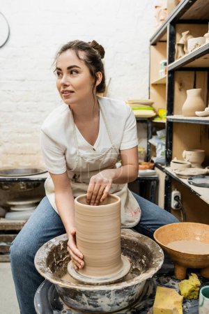 Lächelnde brünette Kunsthandwerkerin in Schürze stellt Tonvase auf Töpferscheibe in Werkstatt her