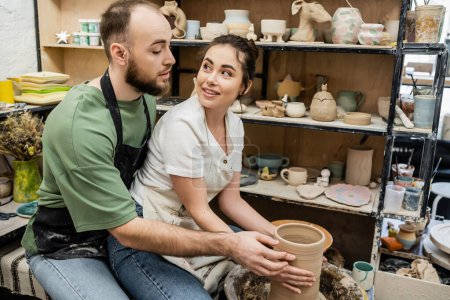 Foto de Artesano sonriente en delantal mirando novio mientras hace jarrón de barro en rueda de cerámica en taller - Imagen libre de derechos