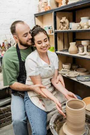 Smiling artisan hugging girlfriend while making clay vase on pottery wheel in ceramic studio
