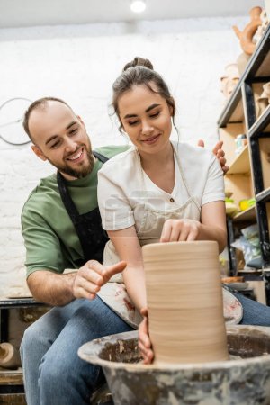 Foto de Artesano sonriente en delantal hablando con su novia haciendo jarrón de barro en rueda de cerámica en el taller - Imagen libre de derechos