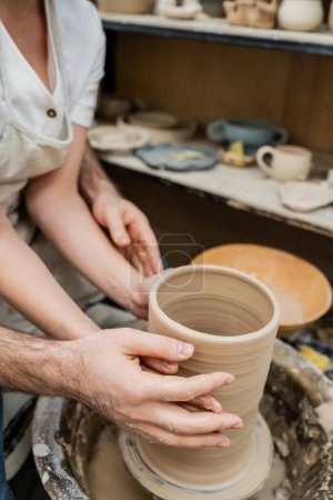 Vista recortada de pareja enamorada haciendo jarrón de arcilla juntos en rueda de cerámica en taller de cerámica