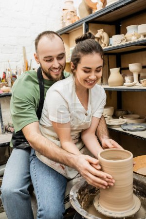 Cheerful craftsman hugging girlfriend and making clay vase together on pottery wheel in workshop