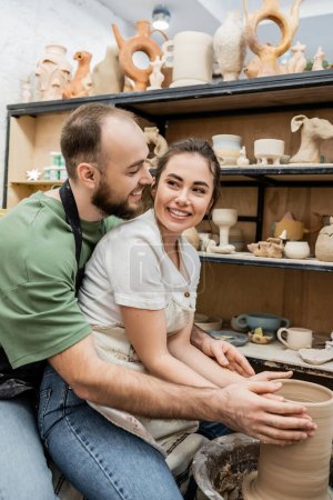 Foto de Artesana positiva en delantal hacer jarrón de arcilla y mirando novio cerca de rueda de cerámica en el estudio - Imagen libre de derechos