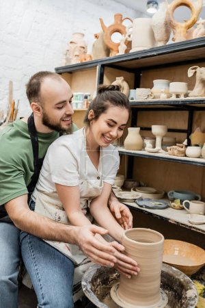 Craftsman in apron shaping clay vase on pottery wheel with smiling girlfriend in ceramic workshop