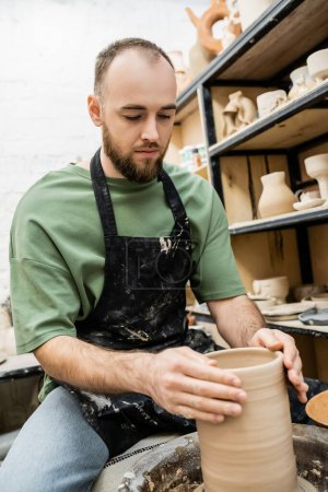 Bearded craftsman in apron shaping clay vase on pottery wheel in ceramic workshop at background