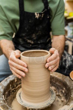 Cropped view of blurred artisan in apron shaping clay vase on pottery wheel in ceramic studio