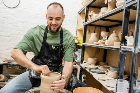 Foto de Alegre barbudo artesano en delantal formando jarrón de arcilla en rueda de cerámica cerca de rack en taller - Imagen libre de derechos