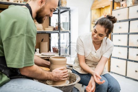 Photo for Smiling craftswoman in apron sitting near boyfriend making clay vase on pottery wheel in workshop - Royalty Free Image