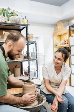 Foto de Alegre artesano en delantal hablando con novio haciendo jarrón de barro sobre rueda de cerámica en taller de cerámica - Imagen libre de derechos