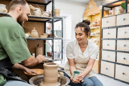 Souriant artisan tenant tasse tandis que petit ami faisant vase d'argile sur roue de poterie en studio