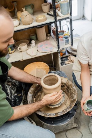 Foto de Vista de ángulo alto de la forma artesanal jarrón de arcilla en la rueda de cerámica cerca de la novia con taza en el estudio - Imagen libre de derechos