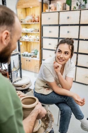 Positive Handwerker in Schürze im Gespräch mit Freundin, die Tonvase auf Töpferscheibe in Werkstatt herstellt