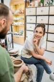 Positive craftsman in apron talking to girlfriend making clay vase on pottery wheel in workshop t-shirt #665331280