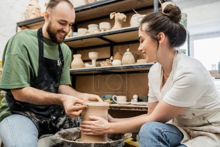 Téléchargez les photos : Artisans romantiques souriants dans des tabliers façonnant vase d'argile ensemble sur la roue de poterie dans l'atelier de céramique - en image libre de droit