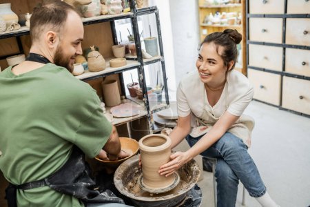 Foto de Artesana sonriente dando forma a jarrón de arcilla en la rueda de cerámica cerca de novio y tazón con agua en el estudio - Imagen libre de derechos