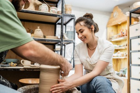 Joyful craftswoman in apron shaping clay vase with blurred boyfriend on pottery wheel in workshop
