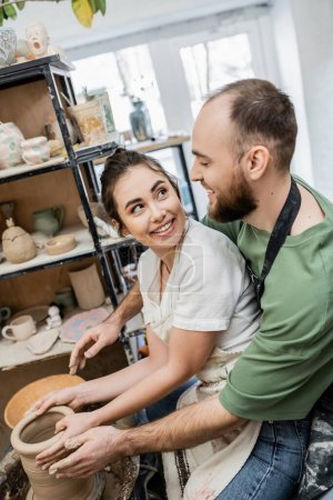 Foto de Artesana alegre en delantal mirando novio y arcilla de moldeo en la rueda de cerámica en el taller - Imagen libre de derechos
