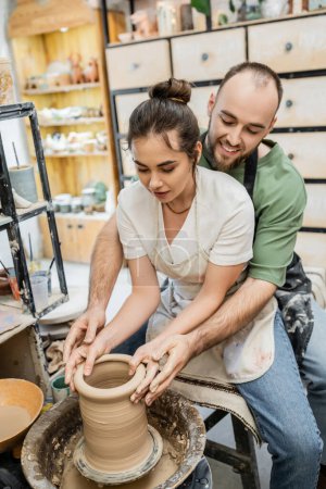 Sonriente barbudo artesano moldeando jarrón de arcilla junto con su novia en la rueda de cerámica en el taller