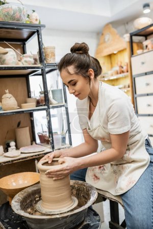 Morena alfarero en delantal la creación de jarrón de arcilla en la rueda de cerámica cerca de tazón con agua en el taller
