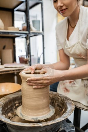 Vista recortada de alfarera femenina en delantal creando jarrón de arcilla en rueda de cerámica en taller borroso