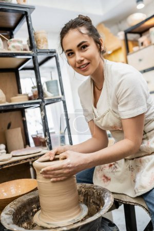 Foto de Alfarero morena sonriente en delantal mirando a la cámara y haciendo jarrón de arcilla en la rueda de cerámica en el estudio - Imagen libre de derechos