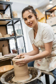 Smiling brunette potter in apron looking at camera and making clay vase on pottery wheel in studio Stickers #665331506