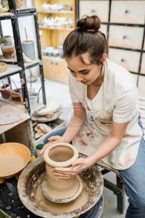 Brünette Kunsthandwerkerin in verschwommener Schürze macht Tonvase auf Töpferscheibe in Wassernähe in Werkstatt