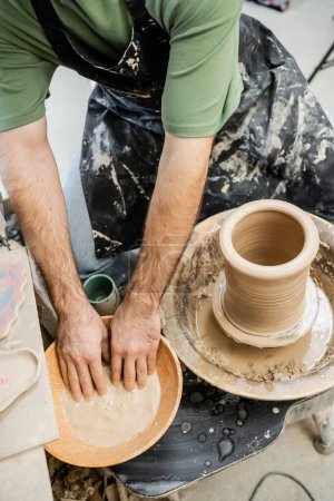Téléchargez les photos : Vue du dessus du potier masculin dans le tablier travaillant avec de l'eau dans un bol et de l'argile sur une roue de poterie en atelier - en image libre de droit