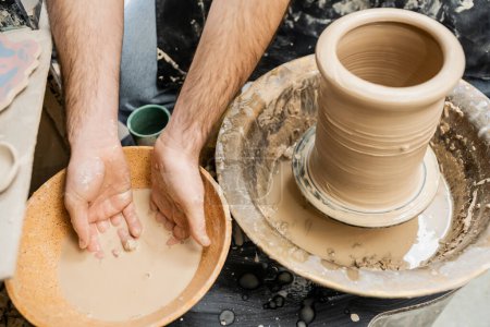Vista superior del artesano en delantal trabajando con agua en tazón y arcilla sobre rueda de cerámica en estudio