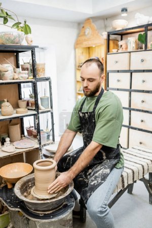 Bearded craftsman in dirty apron shaping and forming clay on pottery wheel in ceramic studio