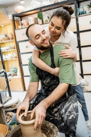 Artesana sonriente en delantal abrazando novio formando arcilla sobre rueda de cerámica en estudio de cerámica