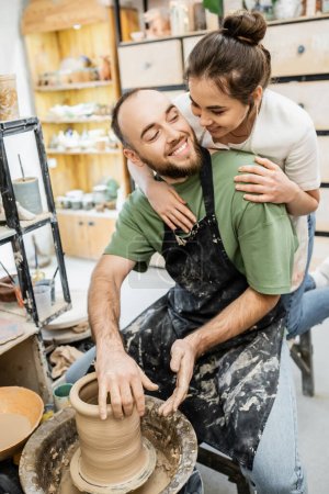 Téléchargez les photos : Artisanat dans tablier embrassant copain souriant façonnant l'argile sur la roue de poterie dans l'atelier - en image libre de droit