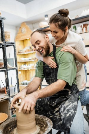 Joyful craftswoman in apron embracing boyfriend making clay vase on pottery wheel in workshop