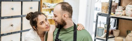 Foto de Morena artesana en delantal abrazando y mirando novio en taller de cerámica, pancarta - Imagen libre de derechos