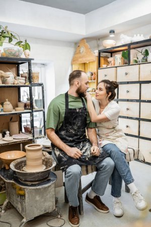 Artesana en delantal abrazando novio y hablando cerca de arcilla sobre rueda de cerámica en taller