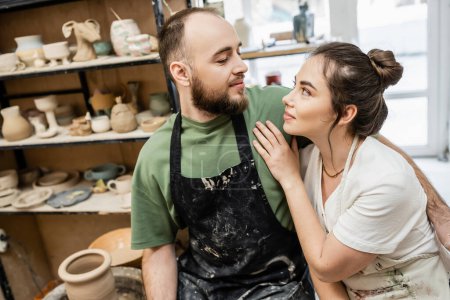 Photo for Bearded craftsman in apron hugging and looking at girlfriend near blurred clay sculptures in studio - Royalty Free Image