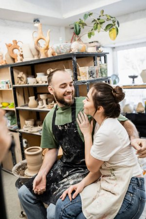 Fröhlicher bärtiger Bildhauer in Schürze umarmt Freundin nahe verschwommener Ton- und Töpferscheibe in Werkstatt