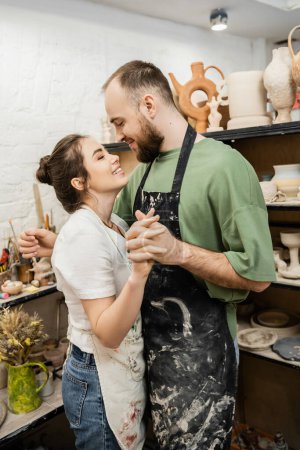 Lächelndes Handwerkerpaar in Schürzen hält Händchen und umarmt sich in der Keramikwerkstatt