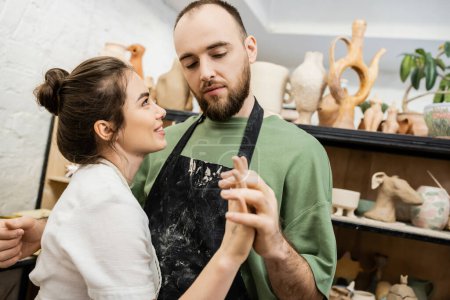 Sculpteur barbu dans un tablier tenant la main d'une petite amie souriante tout en travaillant dans un atelier de céramique