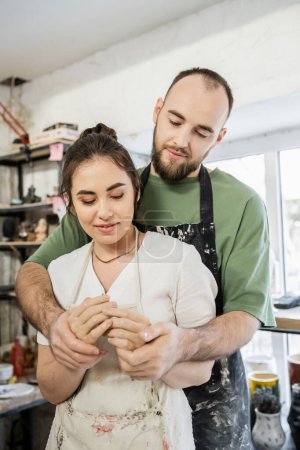 Bearded craftsman holding hands and hugging smiling girlfriend in apron in ceramic workshop