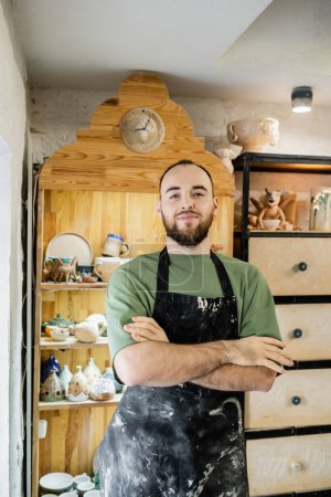 Confident bearded craftsman in apron crossing arms and looking at camera in ceramic workshop
