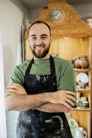 Cheerful bearded craftsman in apron crossing arms and looking at camera while standing in workshop