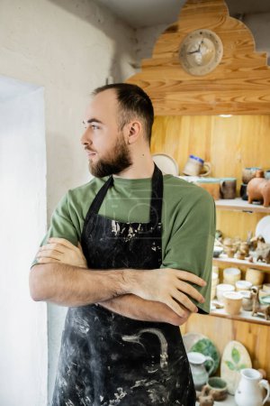 Artisan barbu dans un tablier croisant les bras et regardant loin tout en restant debout dans un atelier de céramique