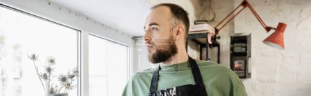 Photo for Bearded sculptor in apron looking away while standing near flowers in ceramic workshop - Royalty Free Image