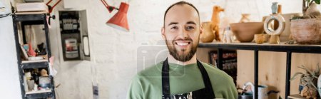 Photo for Smiling craftsman in apron looking at camera and standing in blurred ceramic workshop, banner - Royalty Free Image